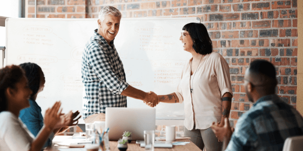 man and a woman stood in front of a white board in a board room shaking hands and smiling